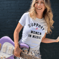 Load image into Gallery viewer, Woman wearing a 'Support Women in Music' t-shirt holding a purple guitar against a black brick wall.