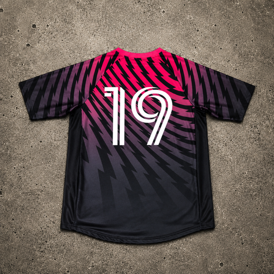 A black soccer jersey with a vibrant pink, purple & black gradient pattern, featuring the number 19 in white on the back. The jersey is laid flat on a textured gray concrete surface.