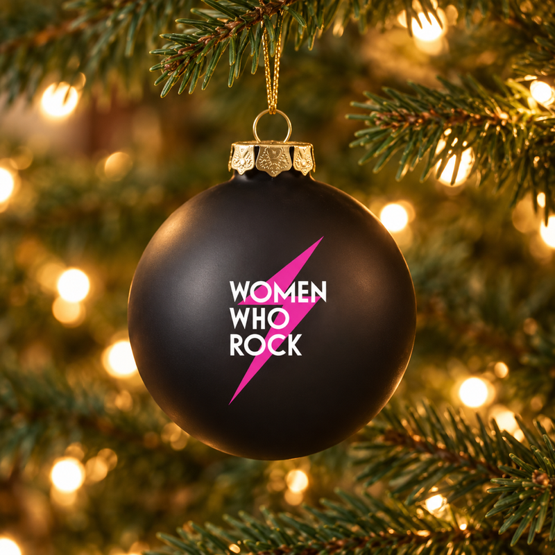 Women Who Rock black glass ornament hanging on a green Christmas tree with warm white lights.