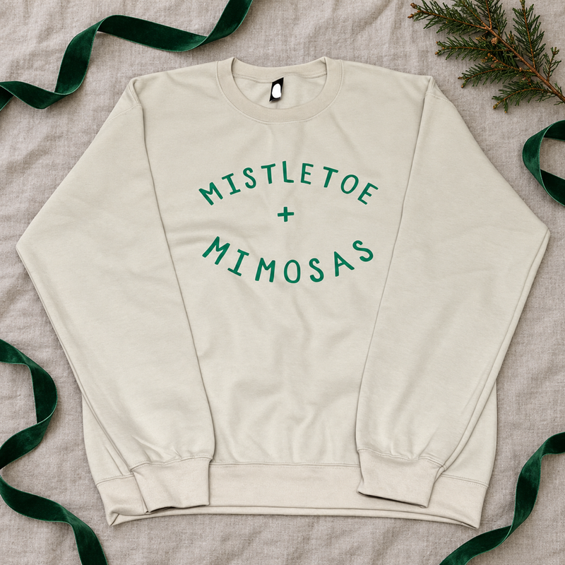Cream Mistletoe + Mimosas crewneck laid flat on linen with green ribbon and evergreen.