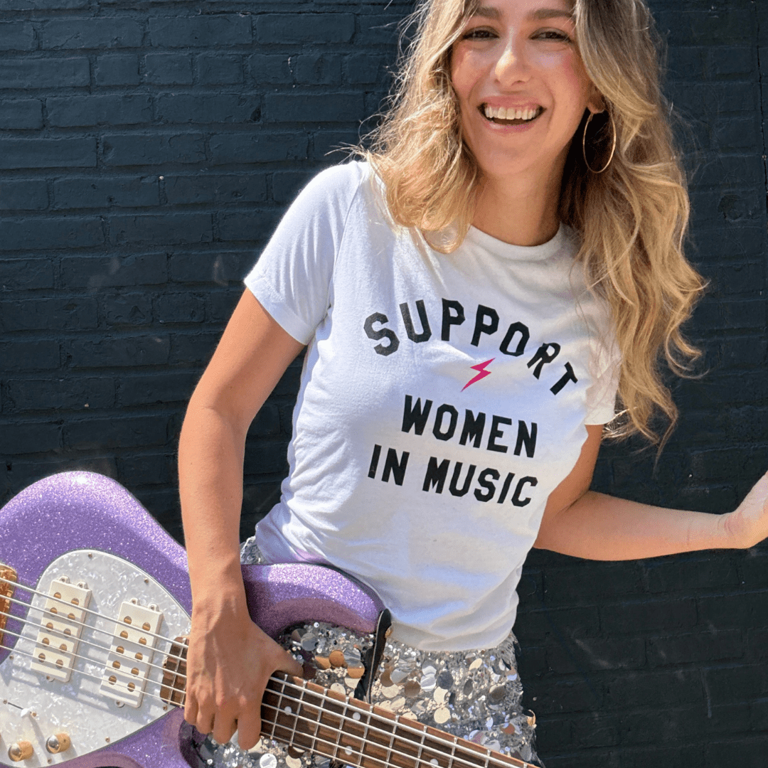 Woman wearing a 'Support Women in Music' t-shirt holding a purple guitar against a black brick wall.