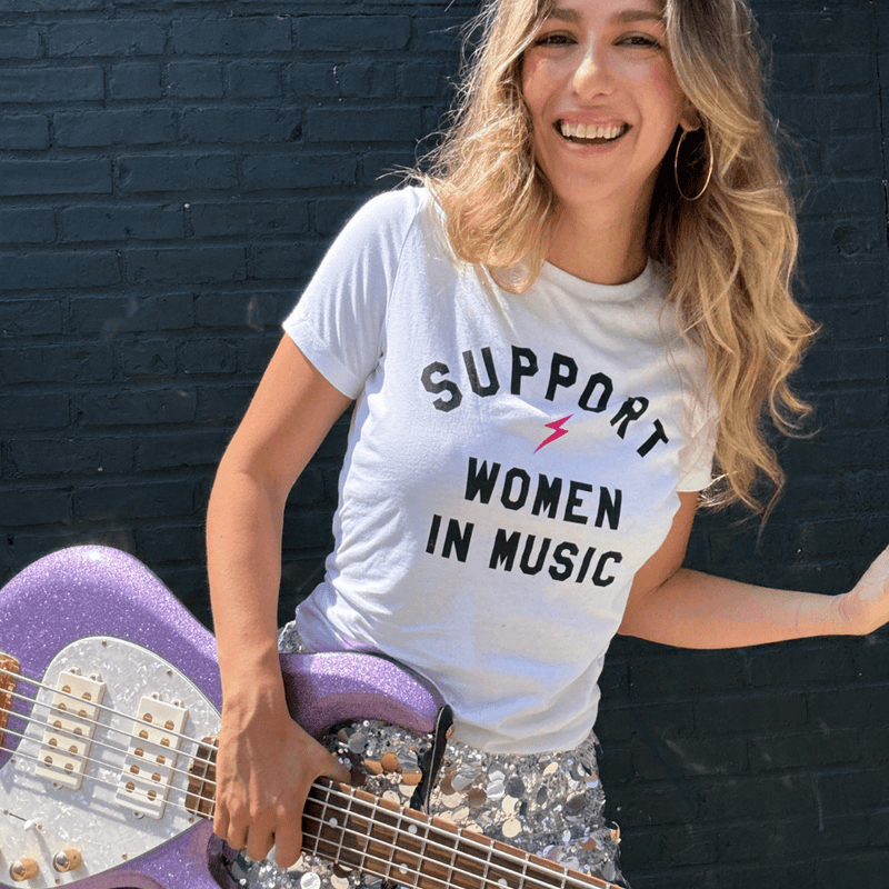 Woman wearing a 'Support Women in Music' t-shirt holding a purple guitar against a black brick wall.
