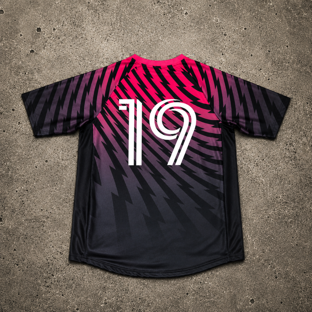 A black soccer jersey with a vibrant pink, purple & black gradient pattern, featuring the number 19 in white on the back. The jersey is laid flat on a textured gray concrete surface. 