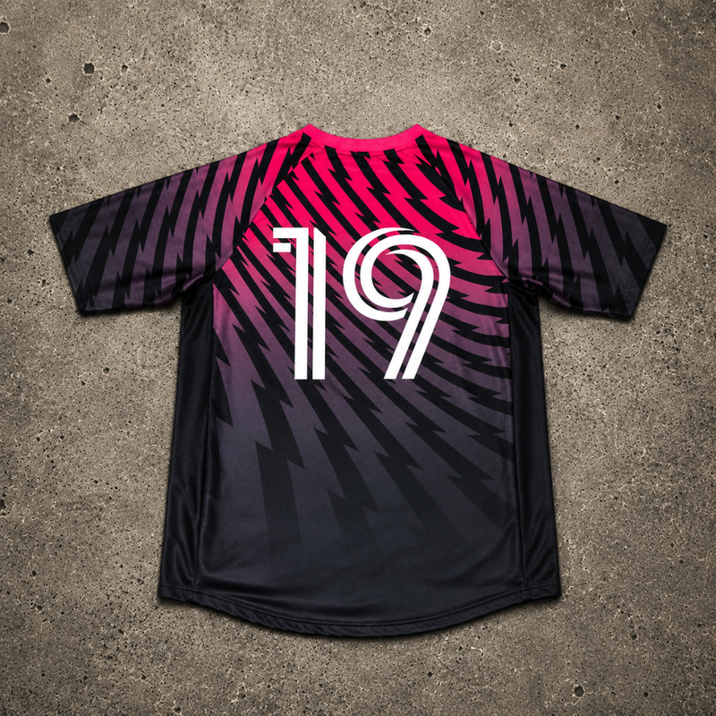 A black soccer jersey with a vibrant pink, purple & black gradient pattern, featuring the number 19 in white on the back. The jersey is laid flat on a textured gray concrete surface. 